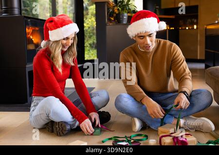 Couple diversifié portant des chapeaux de Père Noël enveloppant des cadeaux dans le salon moderne en utilisant des boîtes et du ruban Banque D'Images