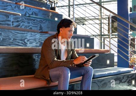 Homme asiatique au milieu des années 20 assis sur un banc à plusieurs étages dans le hall du bureau tenant un ordinateur tablette Banque D'Images