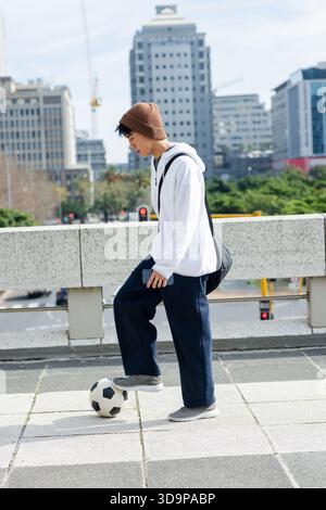Adolescent asiatique mâle en sweat à capuche équilibrant ballon de football tenant smartphone et sac à dos sur le pont Banque D'Images