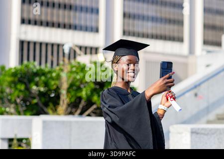 Femme afro-américaine souriante en robe casquette avec diplôme prenant selfie téléphone sur le campus, espace copie Banque D'Images