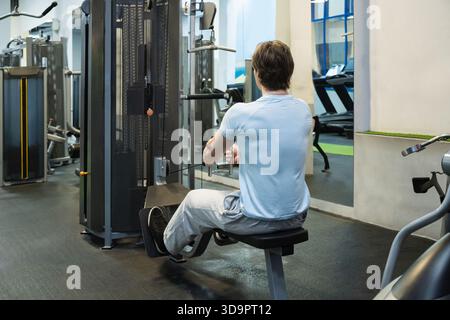 Jeune homme sportif portant un tshirt bleu s'exerçant sur une machine à ramer basse au gymnase. Construction musculaire. Mode de vie sain routine d'entraînement Banque D'Images