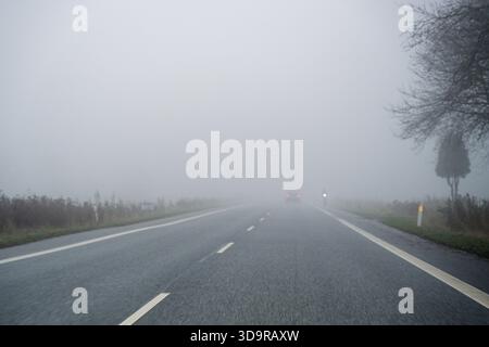 Une voiture se déplace le long d'une route de campagne brumeuse avec une très faible visibilité, créant une atmosphère silencieuse idéale pour des thèmes tels que la sécurité routière, la météo co Banque D'Images