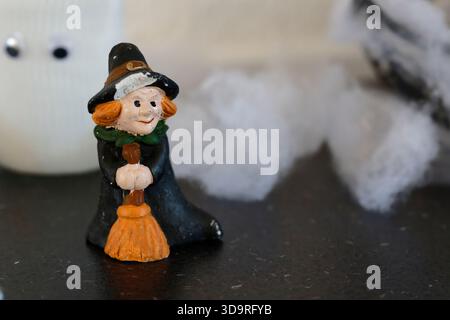 Une petite figure de sorcière en céramique peinte avec des cheveux orange, chapeau noir et manteau tenant un balai, avec un bord de chapeau ébréché, placé sur une table en marbre noir ne Banque D'Images
