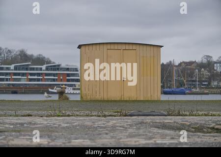 Mur en bois jaune avec des portes près de l'asphalte du port avec mouette assise à proximité et bateaux en arrière-plan par un poste d'amarrage en métal au port de flensburg Banque D'Images