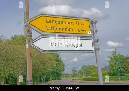 Deux panneaux de signalisation indiquent les directions à gauche pour Langenhorn et Luetjenholm. Le signe blanc indique Materialdepot. Route avec voiture et arbres en arrière-plan. Banque D'Images