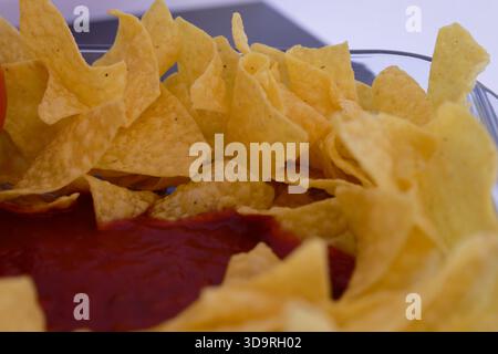 Un bol en verre transparent contient des nachos jaunes croquants disposés autour du bord. Au centre, il y a une sauce rouge, peut-être du ketchup ou une trempette épicée. Banque D'Images