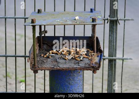 Une petite boîte en métal rouillé est attachée à un poteau bleu à côté d'une clôture métallique. Il est recouvert de mégots de cigarettes, de saleté et même d'un morceau de chewing-gum mâché. Banque D'Images