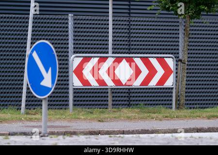 Un panneau de signalisation comportant une série de chevrons rouges indiquant une courbe à l'avant, avec une flèche bleue pointant vers la gauche au premier plan. L'arrière-plan s'affiche Banque D'Images