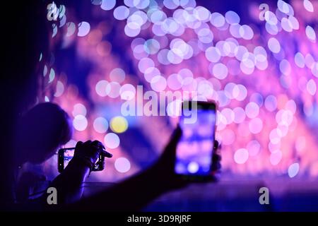 Une foule de personnes capture un spectacle de néons vibrant sur leurs smartphones. Les lumières blanc violet bleu et rose brillent brillamment pour créer une couleur vive Banque D'Images