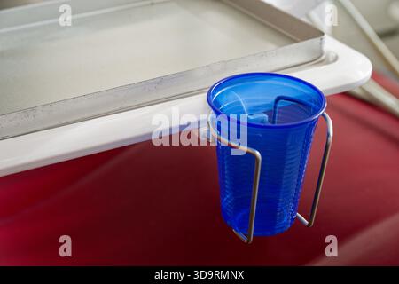 Un support métallique dans une salle de traitement contient une tasse jetable bleue et un plateau en acier inoxydable brillant. Le mur blanc brillant et la disposition ordonnée suggèrent une clinique setti Banque D'Images