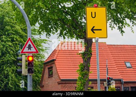 Une intersection de ville montre un feu rouge à côté d'un avertissement de route principale triangulaire et une flèche jaune pointant vers la gauche. Arbres et une maison avec un re Banque D'Images