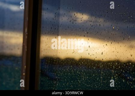 De fines gouttelettes s'accrochent au verre et attrapent la dernière lumière tandis qu'un doux coucher de soleil s'estompe au-delà de la vitre et des toits, les petites perles transforment la vue en un SO Banque D'Images