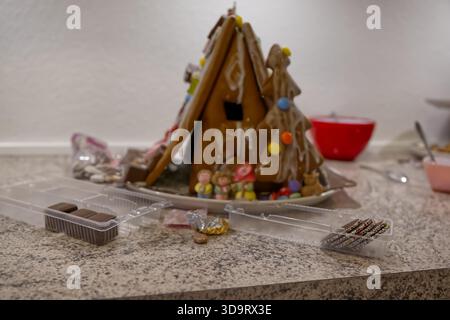 Une maison en pain d'épice décorée avec des bonbons colorés, située sur un comptoir en granit. A proximité se trouvent des friandises au chocolat et un bol rouge, créant un atmos festif Banque D'Images