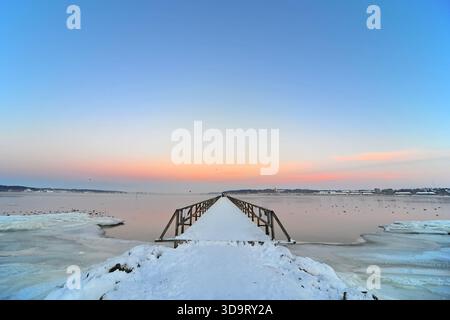 Une scène hivernale sereine avec une jetée en bois s'étendant dans un plan d'eau calme, entouré de glace et de neige. Le ciel est peint avec un col pastel doux Banque D'Images