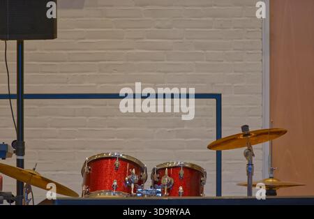 Une vue rapprochée d'un tambour rouge avec des cymbales, positionné contre un mur de briques blanches. La configuration comprend un pied de microphone et une disposition de scène simple Banque D'Images