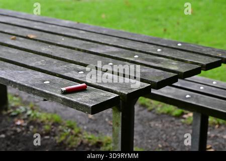 Une vue rapprochée d'une table de pique-nique en bois altérée avec un seul allume-cigare rouge posé dessus. L'arrière-plan présente une pelouse verte luxuriante, creat Banque D'Images