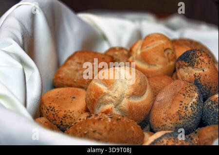 Un panier rempli de différents types de petits pains fraîchement cuits, y compris des variétés de graines de pavot et de graines de sésame, disposés soigneusement sur un chiffon blanc. Banque D'Images