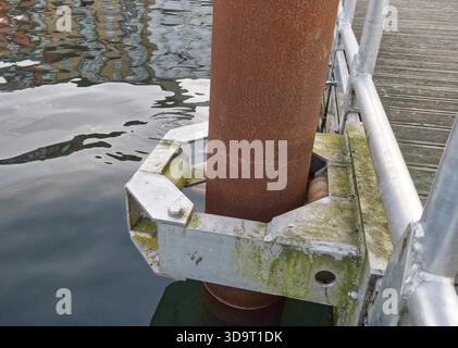 Vue rapprochée d'un poteau en métal rouillé fixé à un quai avec une pince métallique. L'eau reflète les bâtiments environnants, créant une atmosphère sereine Banque D'Images