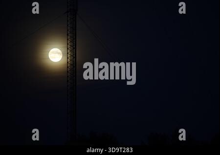 Une pleine lune partiellement obscurcie par des nuages, brillante contre un ciel nocturne sombre. Une grande tour de radio se dresse au premier plan, ajoutant d Banque D'Images