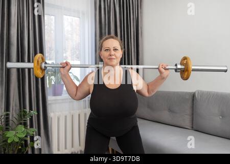 Femme d'âge moyen faisant des squats à barre dans son salon. pour illustrer les routines de fitness à domicile, la musculation et les modes de vie sains. Banque D'Images
