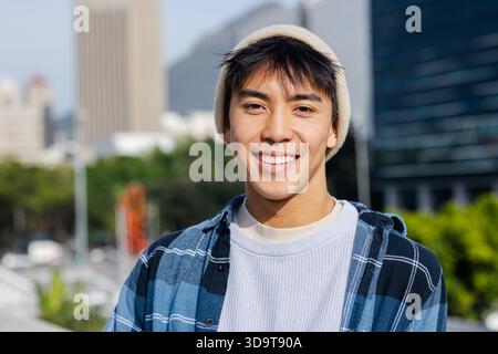 Homme asiatique debout à l'extérieur sur une passerelle urbaine portant un bonnet beige tricoté et une chemise à carreaux bleus Banque D'Images