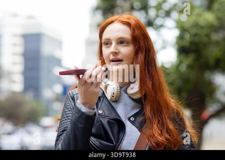 Femme aux cheveux rouges portant une veste en cuir et des écouteurs parlant dans un smartphone sur le trottoir de la ville Banque D'Images