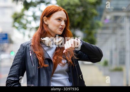 Femme dans ses années 30 regardant smartwatch sur le trottoir tout en portant une veste en cuir et des écouteurs Banque D'Images