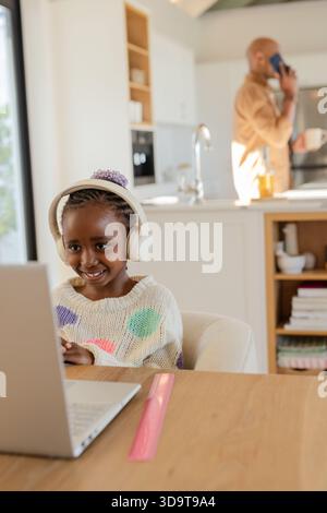 Fille noire d'âge scolaire utilisant un ordinateur portable portant des écouteurs à la table de cuisine à la maison avec règle, espace de copie Banque D'Images