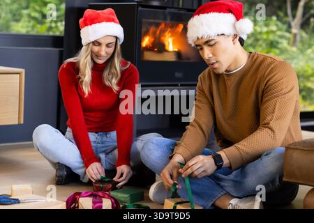 Couple portant des chapeaux de Père Noël assis et enveloppant des cadeaux de Noël par cheminée avec des boîtes rubans ciseaux Banque D'Images