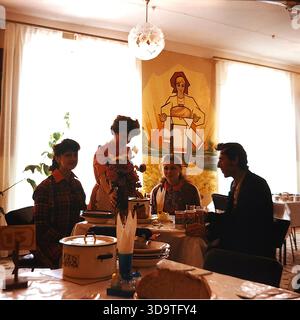 Photographie d'archives vintage des années 1980 capturant une scène de déjeuner au sanatorium Bogatyr à Sloviansk, URSS. Une serveuse dans un tablier sert de la soupe à trois jeunes travailleurs assis à une table avec du kéfir et des fleurs. Le mur de la salle à manger présente une peinture murale représentant une femme en costume traditionnel ukrainien tenant une miche de pain, symbolisant l'hospitalité. Cette image authentique reflète la protection sociale et la vie quotidienne des employés de l'usine Khimprom dans la paisible région du Donbass pendant l'ère soviétique. Banque D'Images