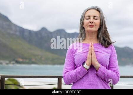 Femme senior debout fermant les yeux pressant les mains sur la poitrine à la garde-corps de pont smartwatch, espace de copie Banque D'Images