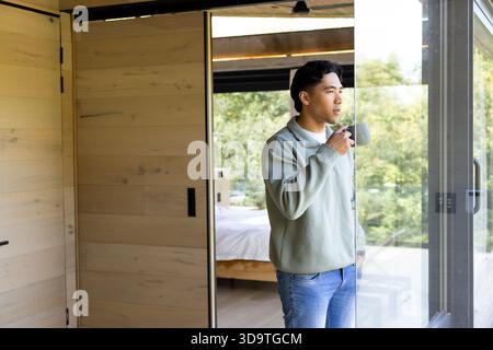 Homme asiatique debout dans la chambre tenant tasse à côté du lit regardant à travers la porte vitrée coulissante, espace de copie Banque D'Images