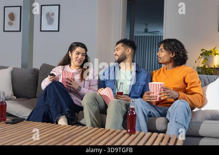 Divers amis regardant le film sur le canapé gris dans le salon tenant des baignoires à pop-corn et des bouteilles de soda Banque D'Images