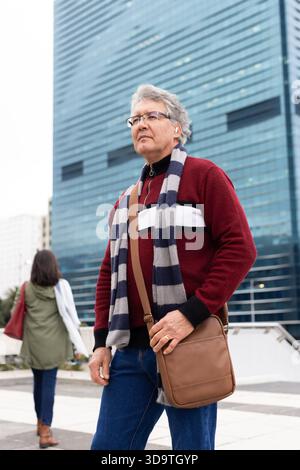 Homme senior debout sur la place de la ville portant un pull rouge et une écharpe grise tenant un sac de messager brun Banque D'Images