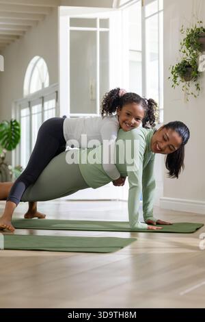 Mère asiatique en vêtements de sport soutenant la fille afro-américaine pendant la planche sur tapis de yoga à la maison Banque D'Images
