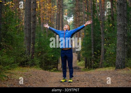 Jeune homme debout sur un sentier forestier avec les bras tendus, respirant l'air frais de l'automne et profitant d'un moment paisible et énergisant de liberté et de pleine conscience tout en faisant du jogging Banque D'Images