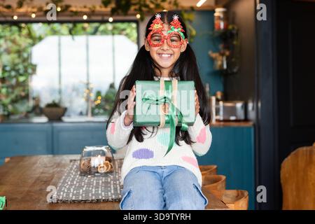 Fille indienne enfant portant des lunettes festives souriant tout en tenant la boîte-cadeau verte à la table de la cuisine Banque D'Images