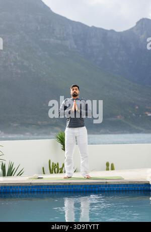 Homme indien en vêtements de sport effectuant la pose de prière sur le tapis de yoga avec des succulentes en pot sur la terrasse de la piscine Banque D'Images