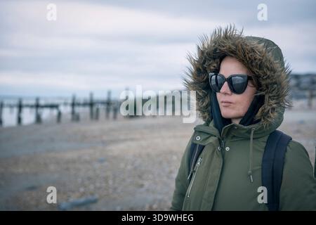Femme en fourrure manteau d'hiver à capuche avec lunettes de soleil et sac à dos sur la plage venteuse Banque D'Images