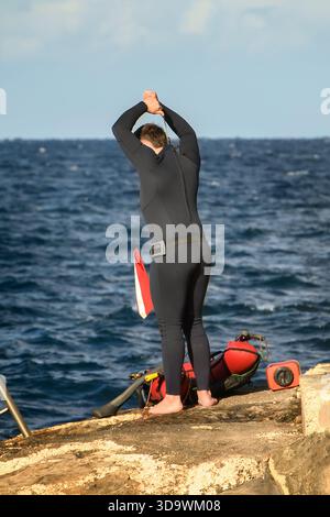 Préparation pour une plongée sous-marine : un plongeur masculin zippe la combinaison noire sur son dos, debout pieds nus sur la rive rocheuse avec son équipement au bord de la mer. Banque D'Images