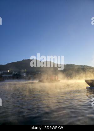 Paysage matinal serein avec lac brumeux au lever du soleil, montrant un brouillard doré s'élevant de l'eau calme. bateau repose dans une belle lumière avec montagne et clair Banque D'Images