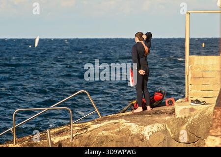 Préparation pour une plongée sous-marine : un plongeur masculin zippe la combinaison noire sur son dos, debout pieds nus sur la rive rocheuse avec son équipement au bord de la mer. Banque D'Images