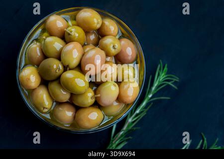 Plan au-dessus d'olives vertes brillantes marinées dans l'huile d'olive, servies dans un bol en verre avec un brin de romarin frais sur un fond texturé foncé. Banque D'Images