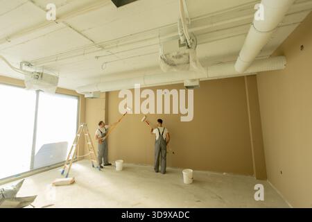 Deux hommes portant une combinaison appliquent de la peinture sur un mur brun à l'aide de rouleaux. Une échelle et des seaux à peinture sont visibles dans la pièce bien éclairée avec un grand vent Banque D'Images
