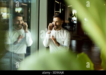 Un homme professionnel en chemise blanche se tient près d'une grande fenêtre, parlant au téléphone. Sa réflexion montre une expression focalisée alors qu'il s'engage dans co Banque D'Images