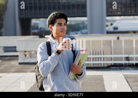 Étudiant masculin debout sur le toit près de la rampe tenant une tasse à café et des cahiers portant un sac à dos Banque D'Images