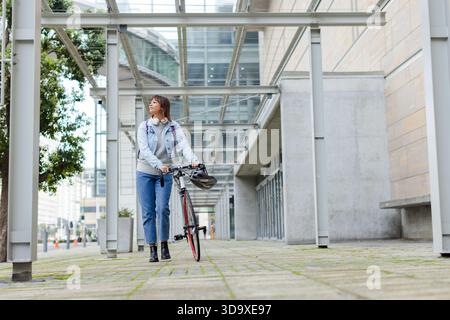 Femme asiatique marchant et poussant le vélo sur la passerelle sous la canopée à la façade du bureau Banque D'Images
