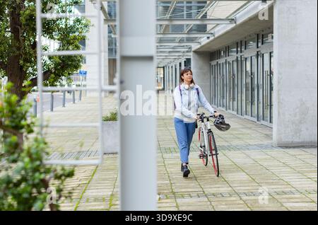 Femme asiatique poussant le vélo sur la plaza en construisant portant veste en denim avec sac à dos et casque Banque D'Images