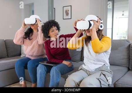 Divers amis féminins assis sur un canapé gris dans le salon en utilisant des casques VR tenant la tablette Banque D'Images