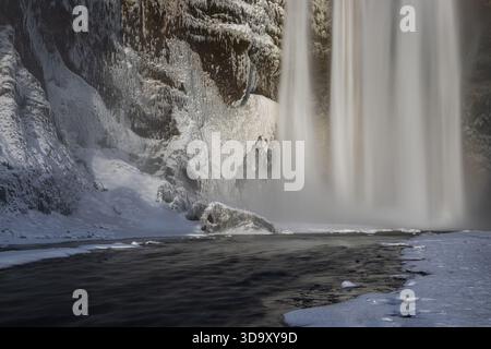 La cascade de Skogafoss coule sur une falaise glacée en Islande pendant l'hiver. Les murs rocheux recouverts de neige et la brume créent une scène contrastée autour de la chute de W. Banque D'Images